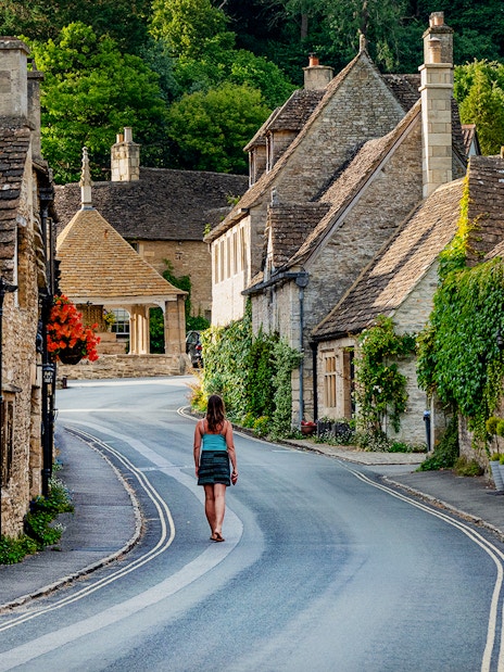 Female tourist walking through a picturesque village in Cotswolds, England, with stone cottages and lush greenery.