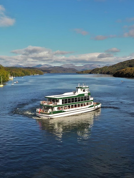 Lake District cruise boat on serene water with scenic hills, part of day tour from London including cream tea.